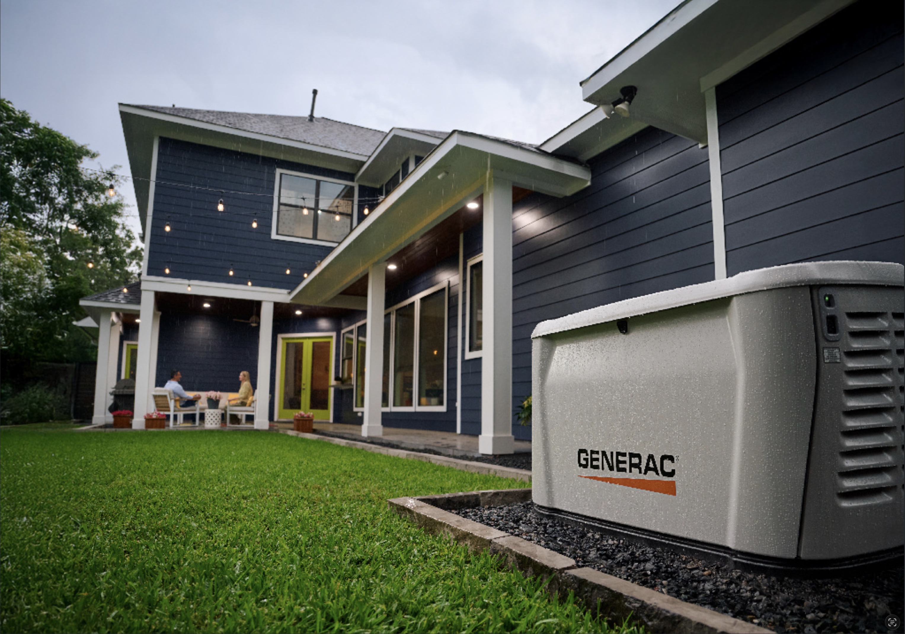 A modern navy home with a lit porch and lawn at dusk, with a standby generator installed alongside